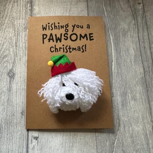 May include: A festive Christmas card featuring a white, fluffy dog wearing a red and green elf hat. The brown card has the text "Wishing you a PAWSOME Christmas!" in black. The dog has a black nose.