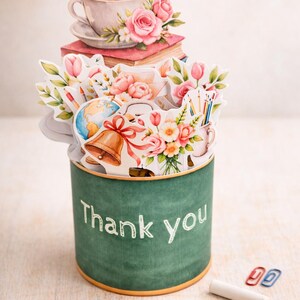 May include: A green cylinder with the words "Thank you" in white script. The cylinder is decorated with paper cutouts of school supplies, flowers, and a teacup. A piece of chalk and paper clips are in the foreground.