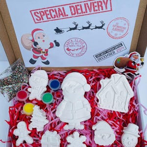 May include: A Christmas craft kit in a cardboard box with red and white tissue paper. The kit includes white ceramic ornaments shaped like Santa, a snowman, a house, a tree, a snowflake, a gift box, and a gingerbread man. The kit also includes paint pots in red, yellow, green, and blue, and a bag of glitter.