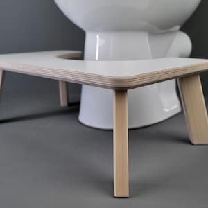 Op de afbeelding: Een witte toiletbrilverhoger van hout met een witte bovenkant. De verhoger is ontworpen om mensen te helpen de toiletbril gemakkelijker te bereiken.