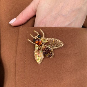 Può includere: Una giacca di tessuto marrone con una spilla a forma di insetto fatta di perline. L'insetto è fatto di perline dorate, marroni e trasparenti e ha una finitura lucida e iridescente.