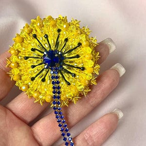 Può includere: Spilla a forma di fiore realizzata a mano con perline gialle, con un centro blu e fili pendenti di strass blu. La spilla presenta un design dettagliato con perline gialle e dettagli neri, creando un accessorio vibrante e accattivante.