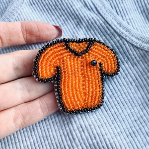 Pode incluir: Broche de contas laranja e preta em forma de camiseta.