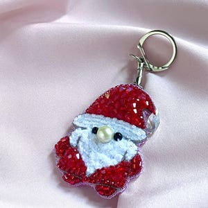 Può includere: Un portachiavi a forma di Babbo Natale realizzato con perline rosse e bianche. La figura presenta un cappello rosso, una barba di perline bianche e un naso a perla centrale. È fissato a un anello in metallo argentato con perline lavorate.