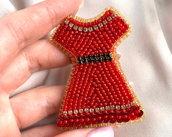 Op de afbeelding: Een rode kralenjurk broche met een zwarte kralenriem en gouden rand. De broche is gemaakt van kleine rode kralen en heeft een gouden omtrek.