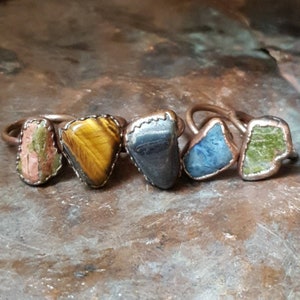 May include: Five rings with different colored stones set in copper. The stones are a pink and green stone, a tiger's eye stone, a dark gray stone, a blue stone, and a green stone.