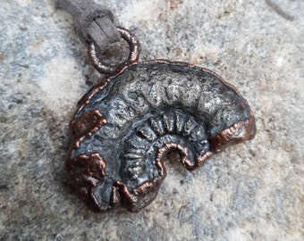 Puede incluir: Un colgante de plata y cobre con forma de hongo con una superficie texturizada. El colgante está unido a un cordón gris.