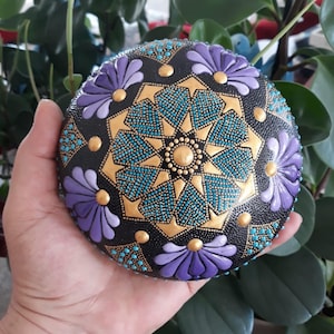 May include: A hand-painted rock with a mandala design. The mandala features a black background with gold and turquoise accents. The center of the mandala is a gold star with a turquoise center. The mandala is surrounded by four purple flowers.