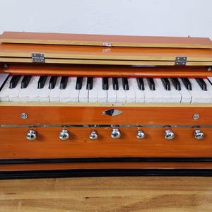 May include: A wooden harmonium with a hinged top and a keyboard of black and white keys. The instrument has a warm, orange-brown finish with silver knobs and decorative metal accents. The harmonium sits on a wooden surface.