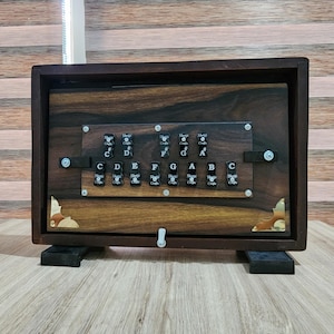 May include: A dark brown wooden musical instrument featuring a rectangular frame with brass corner accents. The front panel displays two rows of black keys labeled with musical notes C, D, E, F, G, A, B, and their sharps, marked Shriji Crafts.