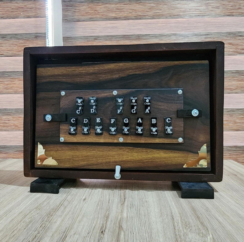 May include: A dark brown wooden musical instrument featuring a rectangular frame with brass corner accents. The front panel displays two rows of black keys labeled with musical notes C, D, E, F, G, A, B, and their sharps, marked Shriji Crafts.