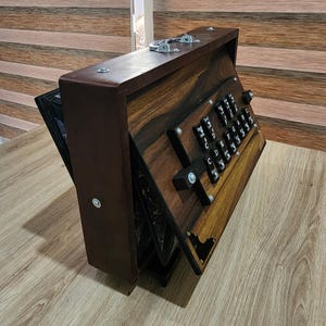 May include: A portable harmonium musical instrument featuring a dark brown wooden casing with a visible grain pattern. The front panel displays a set of black keys labeled with white musical notes including C, D, E, F, G, A, and B, secured by metal hardware.