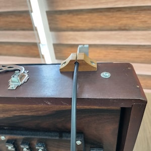 May include: A dark brown wooden box with a visible wood grain, featuring a silver metal latch on the left side and a small wooden cable guide mounted on the top edge. A thin black cable runs through the center of the wooden guide.