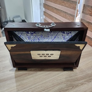 May include: A dark brown wooden harmonium with a rectangular shape, featuring a silver metal handle on top, brass corner accents, and a central metal plate with seven circular cutouts. The interior bellows display a blue and white marbled pattern.
