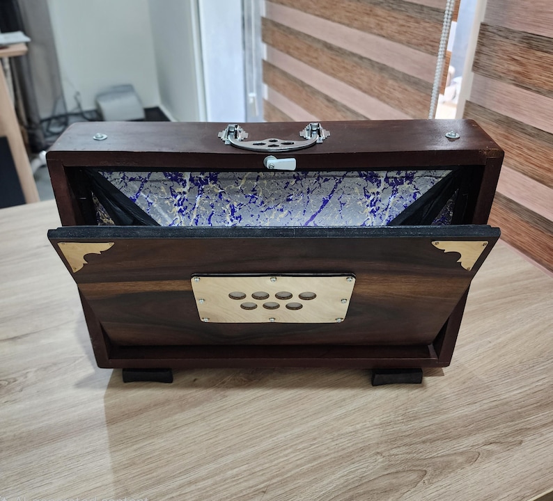 May include: A dark brown wooden harmonium with a rectangular shape, featuring a silver metal handle on top, brass corner accents, and a central metal plate with seven circular cutouts. The interior bellows display a blue and white marbled pattern.