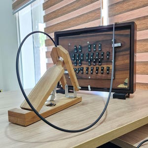 May include: A wooden musical instrument controller featuring a rectangular dark wood frame with a grid of black keys labelled with musical notes. A light wood pedal mechanism with a black cable is positioned in front of the main unit.