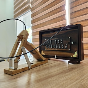 May include: A wooden musical instrument with a dark brown rectangular body featuring black keys labeled with musical notes C, D, E, F, G, A, B, C, paired with a light-colored wooden foot pedal mechanism connected by a black cable.