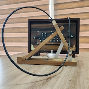 May include: A wooden stand with a triangular frame, a metal spring, and a large black circular hoop, positioned in front of a dark brown wooden musical instrument featuring a series of black keys labeled with musical notes C, D, E, G, A, and C.