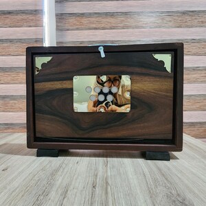 May include: A rectangular dark brown wooden box with a prominent wood grain pattern. The front features a central rectangular metallic plate with seven circular cutouts and gold-toned corner accents. A small white latch is visible at the top center.
