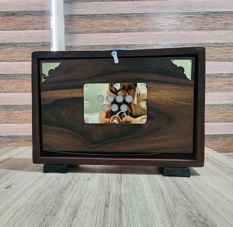 May include: A rectangular dark brown wooden box with a prominent wood grain pattern. The front features a central rectangular metallic plate with seven circular cutouts and gold-toned corner accents. A small white latch is visible at the top center.