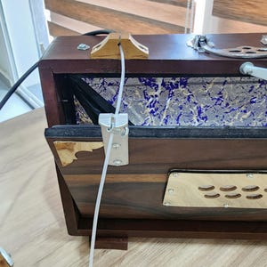 May include: A dark brown wooden harmonium featuring a metallic silver handle, brass corner accents, and a light-colored wooden panel with circular cutouts. The instrument includes a blue and white marbled interior lining and a white cable attachment.
