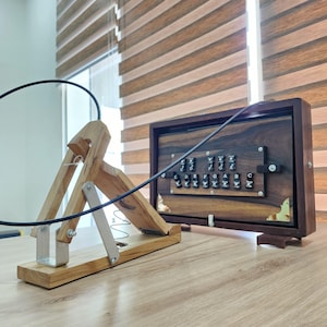 May include: A wooden foot pedal mechanism connected by a black cable to a rectangular wooden harmonium box. The box features a dark wood finish, metal corner accents, and a row of black keys labeled with musical notes C, D, E, F, G, A, B, C.