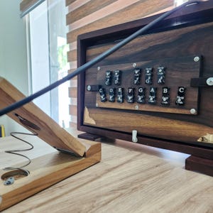 May include: A dark wood harmonium instrument featuring a horizontal panel with black keys labeled with musical notes C, D, E, F, G, A, B, and sharps. A light wood bellows mechanism with a metal spring is positioned in the foreground.