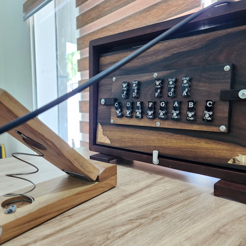 May include: A dark wood harmonium instrument featuring a horizontal panel with black keys labeled with musical notes C, D, E, F, G, A, B, and sharps. A light wood bellows mechanism with a metal spring is positioned in the foreground.