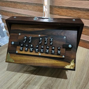 May include: A dark brown wooden harmonium instrument featuring a polished finish, a silver metal handle on top, and gold-toned corner accents. The front panel displays black keys labeled with musical notes C, D, E, F, G, A, B, C, C#, F#, G#.