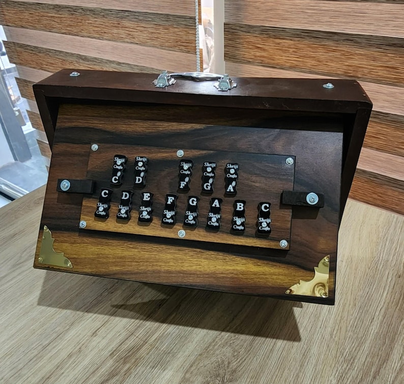 May include: A dark brown wooden harmonium instrument featuring a polished finish, a silver metal handle on top, and gold-toned corner accents. The front panel displays black keys labeled with musical notes C, D, E, F, G, A, B, C, C#, F#, G#.