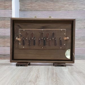 May include: A rectangular wooden musical instrument with a dark brown finish, featuring a central panel with multiple vertical dark keys. The instrument includes brass hardware, a small circular gold-colored accent, and two small wooden feet.