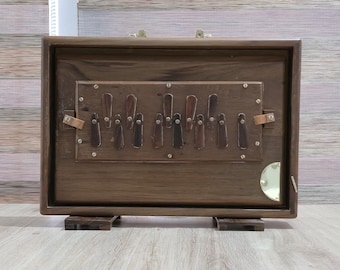 May include: A rectangular wooden musical instrument with a dark brown finish, featuring a central panel with multiple vertical dark keys. The instrument includes brass hardware, a small circular gold-colored accent, and two small wooden feet.