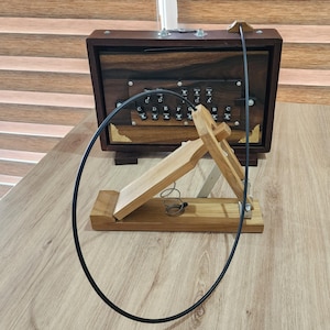 May include: A dark brown wooden harmonium with a black control panel featuring white lettered keys labeled C, D, E, F, G, A, B, and C. A light brown wooden foot pedal with a metal spring is connected to the harmonium by a long black cable.