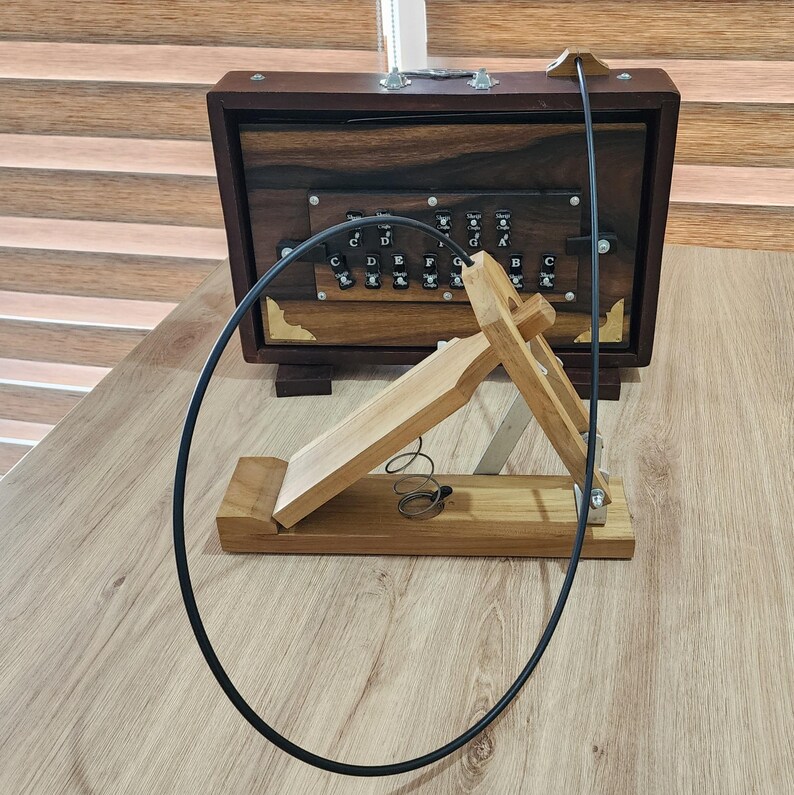 May include: A dark brown wooden harmonium with a black control panel featuring white lettered keys labeled C, D, E, F, G, A, B, and C. A light brown wooden foot pedal with a metal spring is connected to the harmonium by a long black cable.