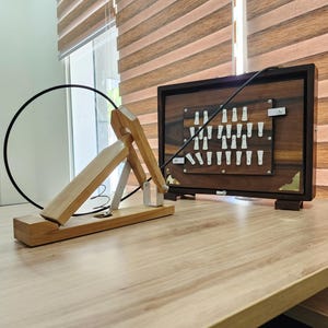 May include: A wooden telegraph key device with a black cable connected to a rectangular wooden box featuring a grid of white plastic components and brass corner accents. The telegraph key has a light wood base and a hinged arm mechanism.