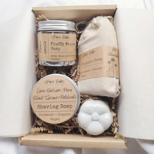 Könnte beinhalten: Eine Geschenkbox mit natürlichen Hautpflegeprodukten. Enthält ein Glas "Fluffy Whipped Soap", eine Dose "Shaving Soap", einen kleinen Beutel "Organic Soap" und eine blütenförmige Badekugel. Die Box ist mit braunem Papier gefüllt.