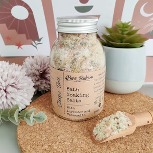 May include: A clear glass jar with a silver lid filled with bath soaking salts. The label reads "Pure Bubs Bath Soaking Salts With Lavender and Chamomile." A small wooden scoop holds a sample of the salts. The jar is on a cork surface.