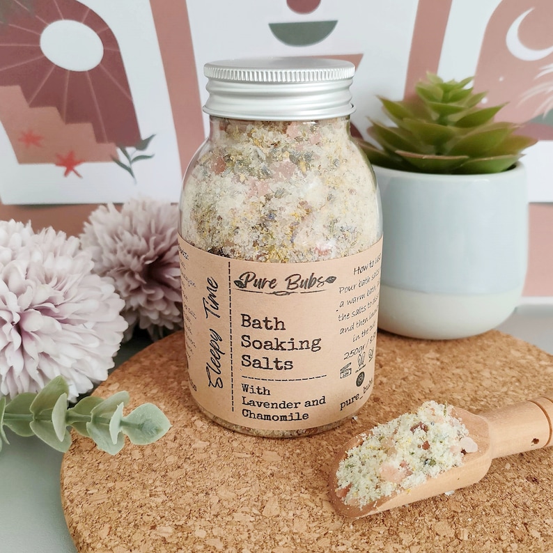 May include: A clear glass jar with a silver lid filled with bath soaking salts. The label reads "Pure Bubs Bath Soaking Salts With Lavender and Chamomile." A small wooden scoop holds a sample of the salts. The jar is on a cork surface.