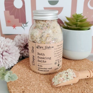 May include: A clear glass jar with a silver lid filled with bath soaking salts. The label reads "Pure Bubs Bath Soaking Salts With Lavender and Chamomile." A wooden scoop holds a small pile of the salts. The jar is on a cork coaster.