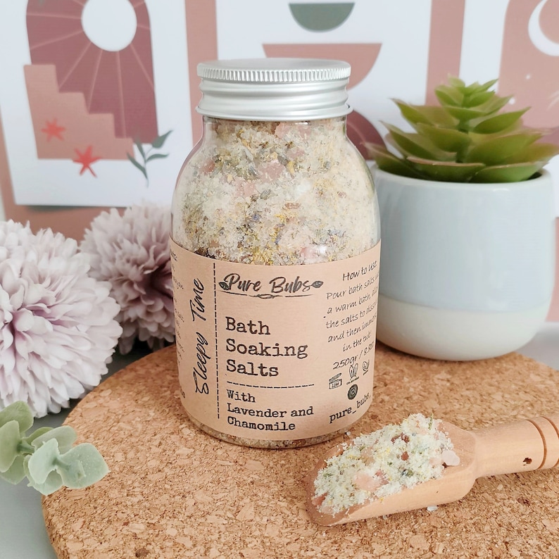 May include: A clear glass jar with a silver lid filled with bath soaking salts. The label reads "Pure Bubs Bath Soaking Salts With Lavender and Chamomile." A wooden scoop holds a small pile of the salts. The jar is on a cork coaster.