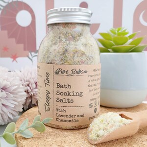 May include: A clear glass jar of bath soaking salts with a silver lid. The label reads "Pure Bubs Bath Soaking Salts with Lavender and Chamomile." The jar contains a mixture of salts and dried herbs. A wooden scoop holds additional salts. A small succulent plant is in the background.