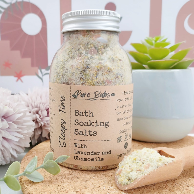 May include: A clear glass jar of bath soaking salts with a silver lid. The label reads "Pure Bubs Bath Soaking Salts with Lavender and Chamomile." The jar contains a mixture of salts and dried herbs. A wooden scoop holds additional salts. A small succulent plant is in the background.