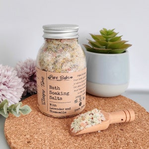 May include: A glass jar with a silver lid contains bath soaking salts. The label reads "Pure Bubs Bath Soaking Salts With Lavender and Chamomile." A wooden scoop holds a small amount of the salts. A small succulent plant in a blue and white pot is in the background.