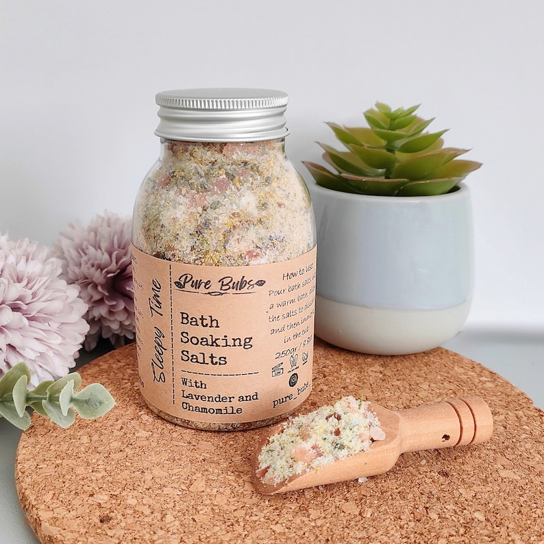 May include: A glass jar with a silver lid contains bath soaking salts. The label reads "Pure Bubs Bath Soaking Salts With Lavender and Chamomile." A wooden scoop holds a small amount of the salts. A small succulent plant in a blue and white pot is in the background.