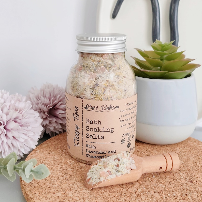 May include: A clear glass jar with a silver lid filled with bath soaking salts. The label reads "Pure Bubs Bath Soaking Salts With Lavender and Chamomile." A small wooden scoop holds a portion of the salts. A small succulent plant is in a white pot.