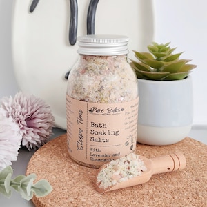 May include: A clear glass jar with a silver lid filled with bath soaking salts. The label reads "Sleepy Time" and "Pure Bubs Bath Soaking Salts With Lavender and Chamomile". A small wooden scoop holds a portion of the salts. A small succulent plant is in a white pot.