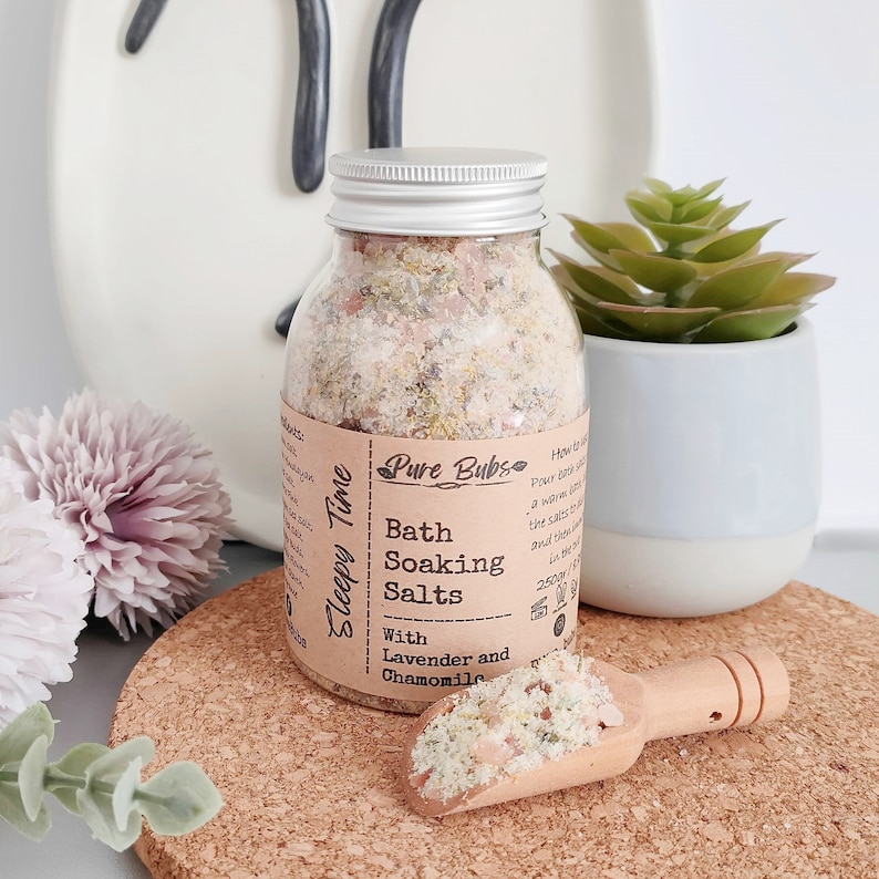 May include: A clear glass jar with a silver lid filled with bath soaking salts. The label reads "Sleepy Time" and "Pure Bubs Bath Soaking Salts With Lavender and Chamomile". A small wooden scoop holds a portion of the salts. A small succulent plant is in a white pot.