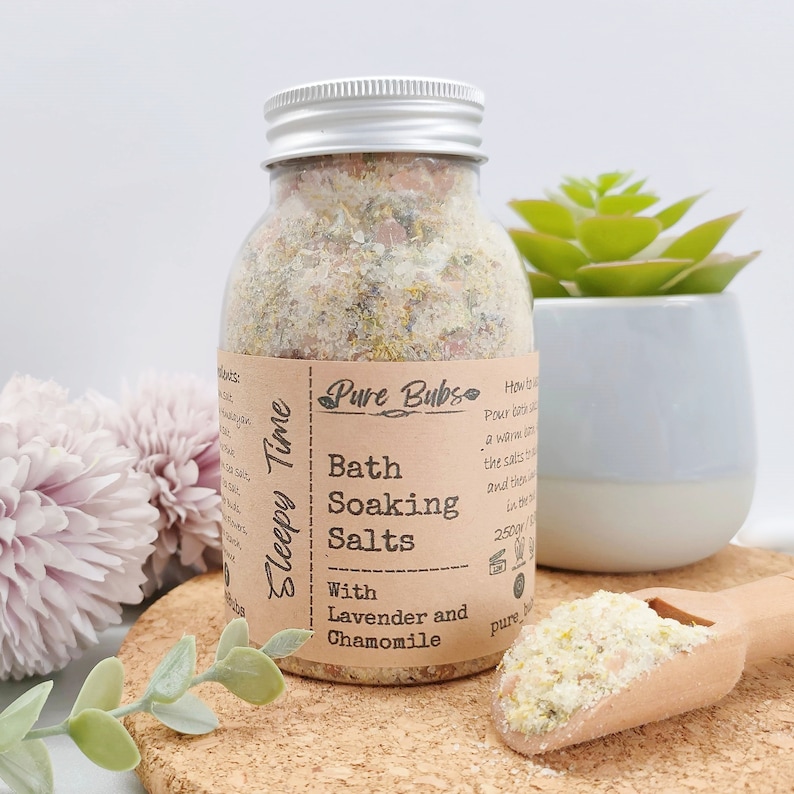 May include: A clear glass jar with a silver lid filled with bath soaking salts. The label reads "Pure Bubs Bath Soaking Salts With Lavender and Chamomile." A wooden scoop holds a small pile of the salts. A small succulent plant sits in a white and blue pot.