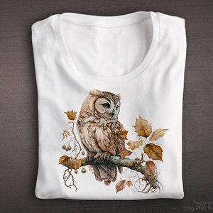 May include: A white t-shirt with a brown and white owl perched on a branch with brown leaves. The owl has large, round eyes and a detailed, realistic design.