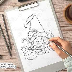 Op de afbeelding: Een zwart-witte printbare kleurplaat met een kerstgnome met een grote hoed, een cadeau en een snoepstok. De tekst "Print thuis en geniet van het kleuren!" staat onderaan de pagina.